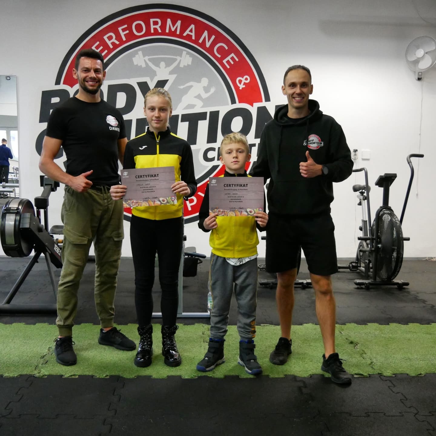 Dwaj uśmiechnięci mężczyźni, prawdopodobnie trenerzy, stoją obok dwójki dzieci trzymających certyfikaty na tle siłowni z logo 'Body Condition Performance & Nutrition Club'.
