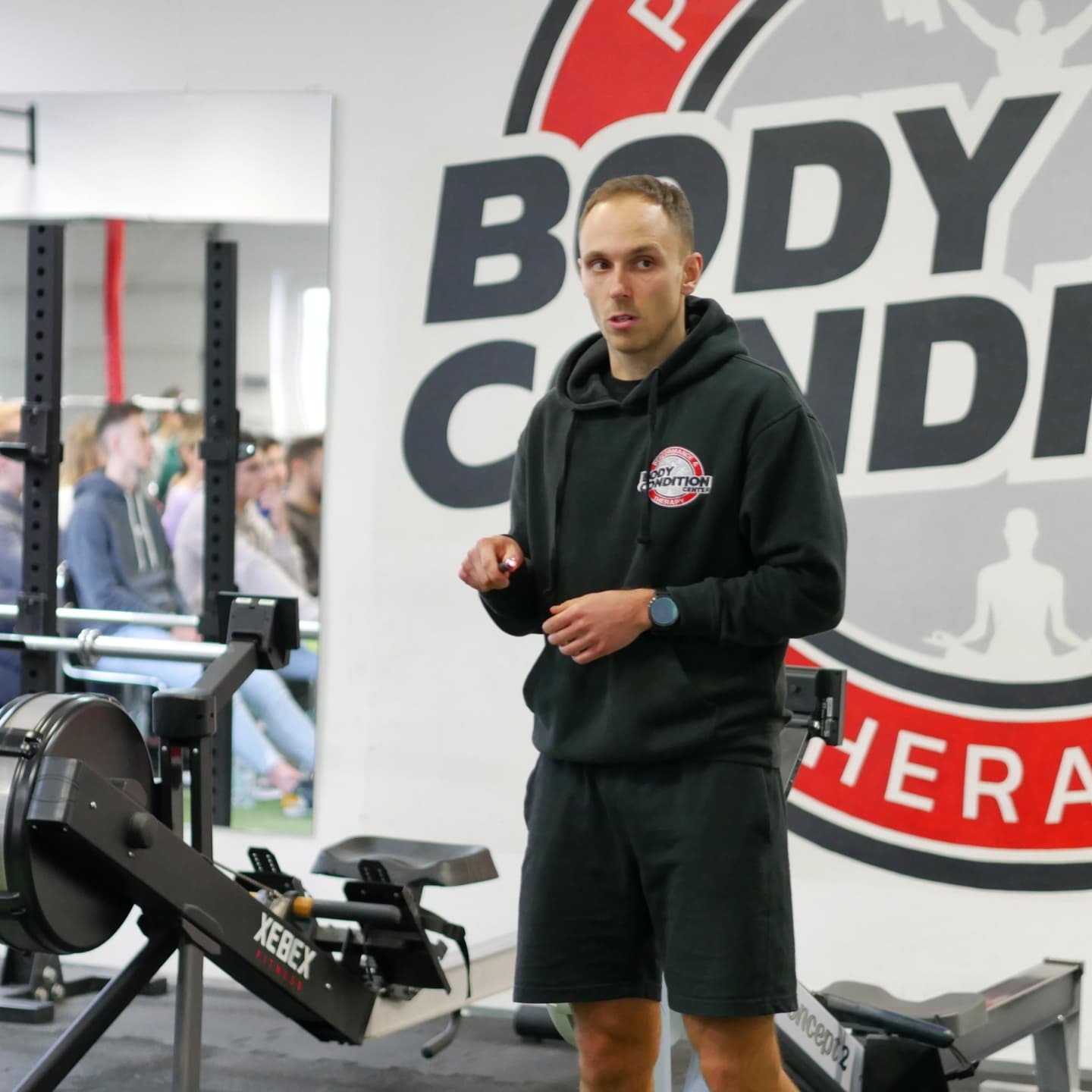 Mężczyzna w czarnej bluzie z kapturem prezentuje ćwiczenie na ergometrze wioślarskim Xebex w siłowni, w tle grupa osób i logo 'Body Condition'.
