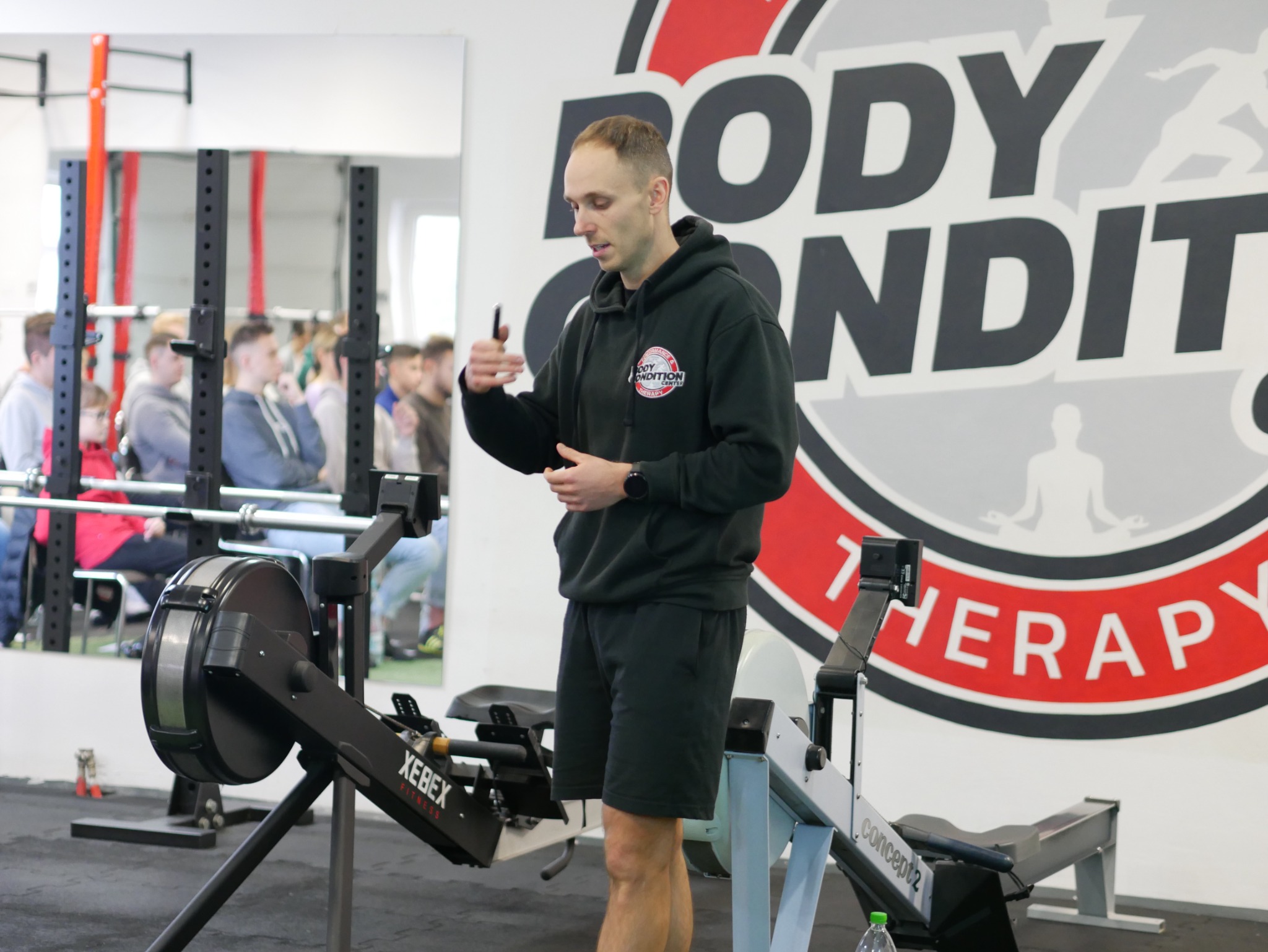 Mężczyzna w czarnej bluzie z kapturem i krótkich spodenkach prezentuje ćwiczenie na ergometrze wioślarskim w siłowni, za nim widoczni obserwatorzy i logo 'Body Condition Therapy'.