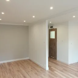 Jasne, przestronne wnętrze po remoncie w Suwałkach: ściany pomalowane na szaro i biało, drewnopodobna podłoga, białe listwy przypodłogowe, drewniane drzwi z szybą i punktowe oświetlenie sufitowe.