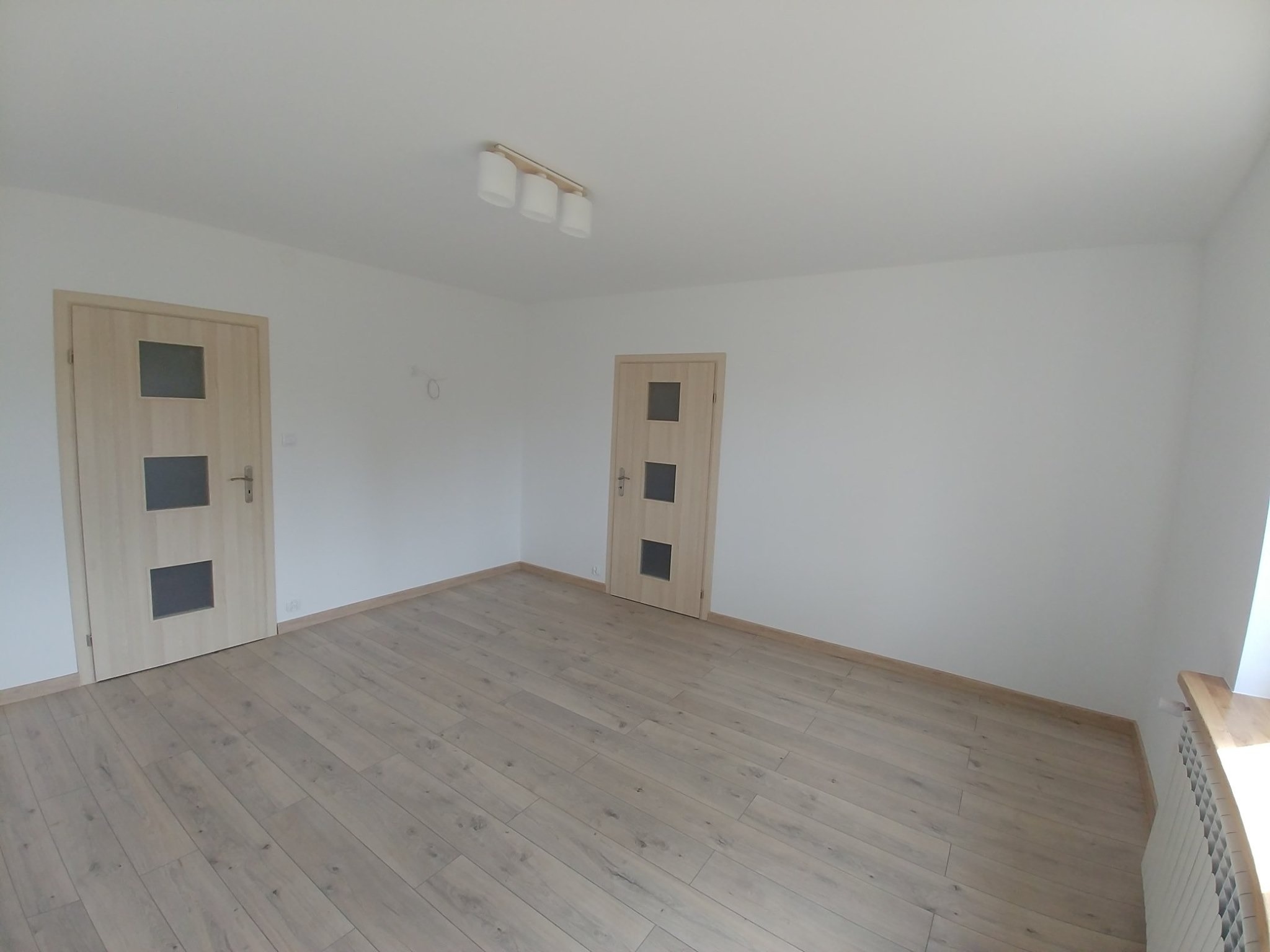 Pusty pokój po remoncie z dwiema jasnymi, drewnianymi drzwiami z szybkami, panelami podłogowymi w odcieniach szarości i białą lampą sufitową z trzema kloszami.