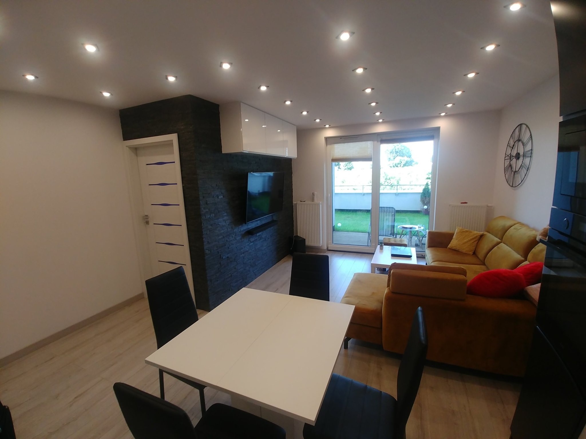 Nowoczesne wnętrze salonu z żółtą sofą, czarną ścianą z kamienia dekoracyjnego, białym stołem i oświetleniem punktowym na suficie.