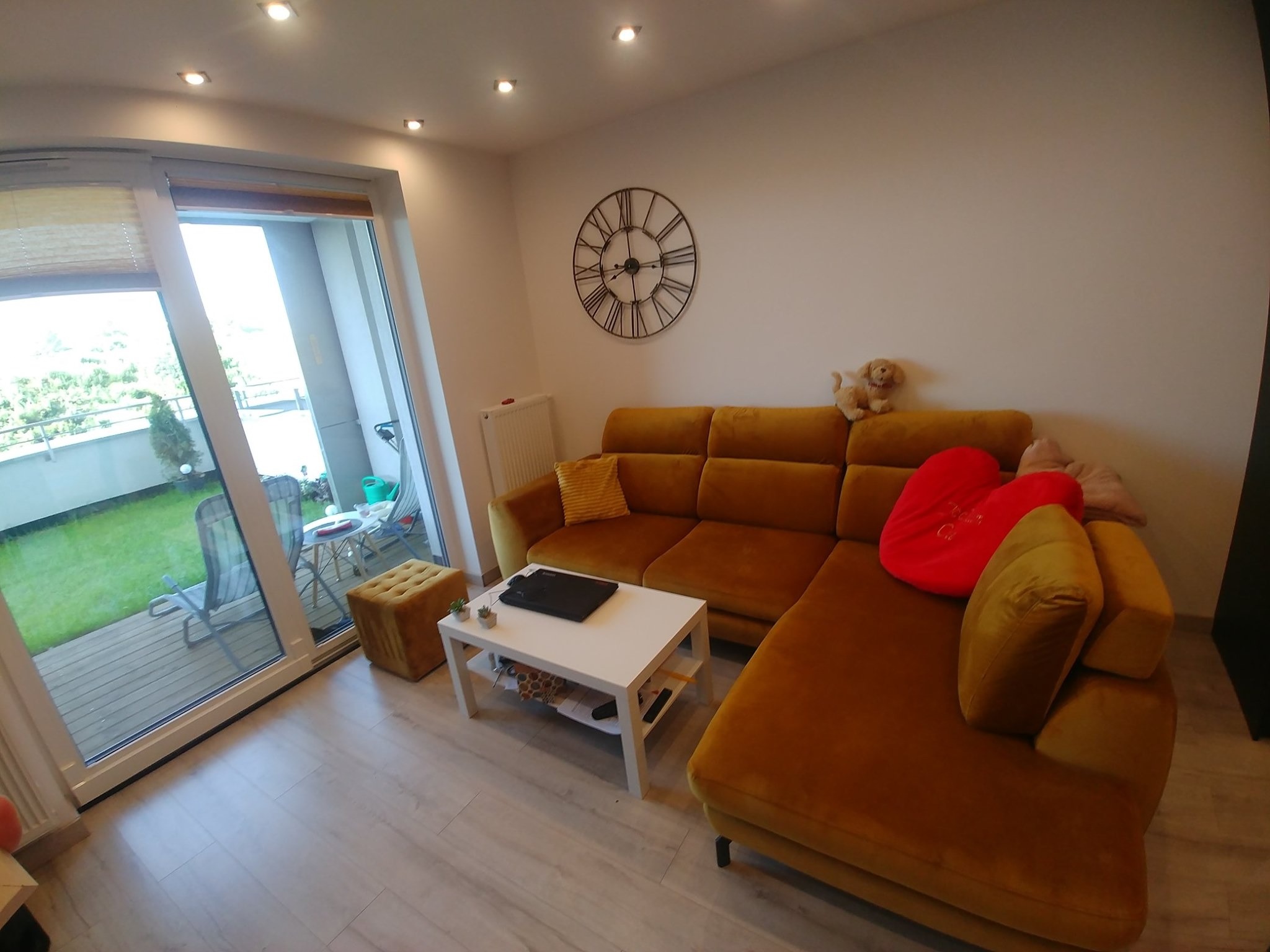 Umeblowane wnętrze pokoju dziennego z narożną sofą w odcieniach musztardowych, białym stolikiem kawowym i widokiem na balkon z zielenią na zewnątrz.