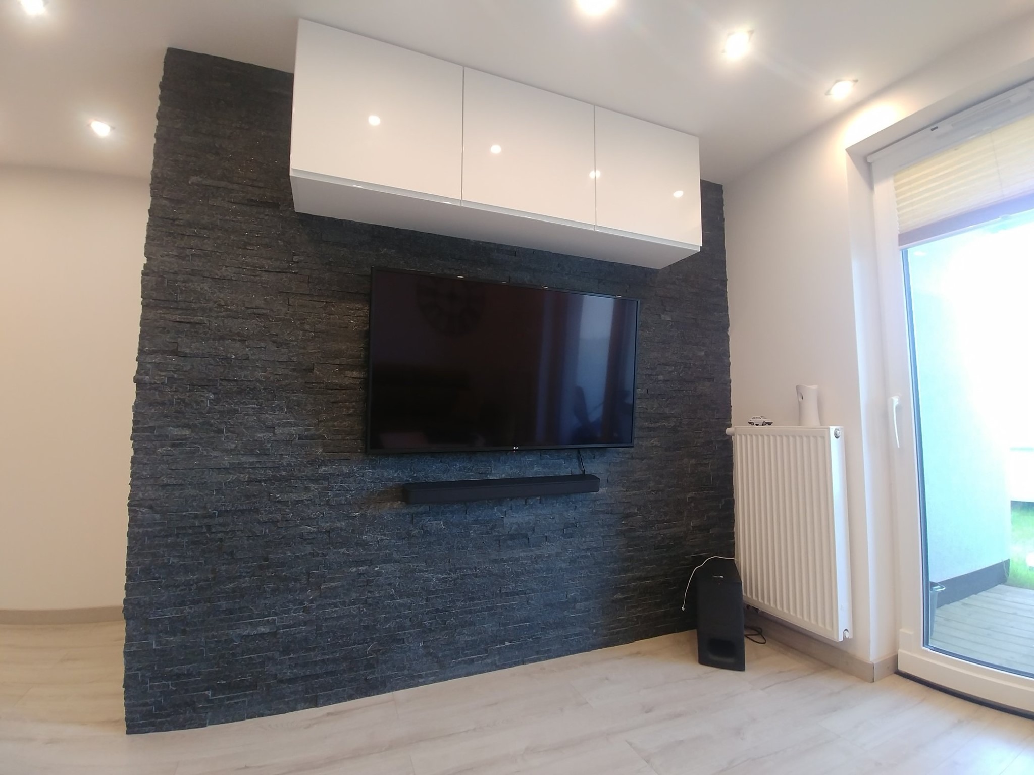 Nowoczesny salon z ścianą wyłożoną ciemnym kamieniem dekoracyjnym, podwieszanymi białymi szafkami, telewizorem, soundbarem, grzejnikiem i oknem balkonowym.