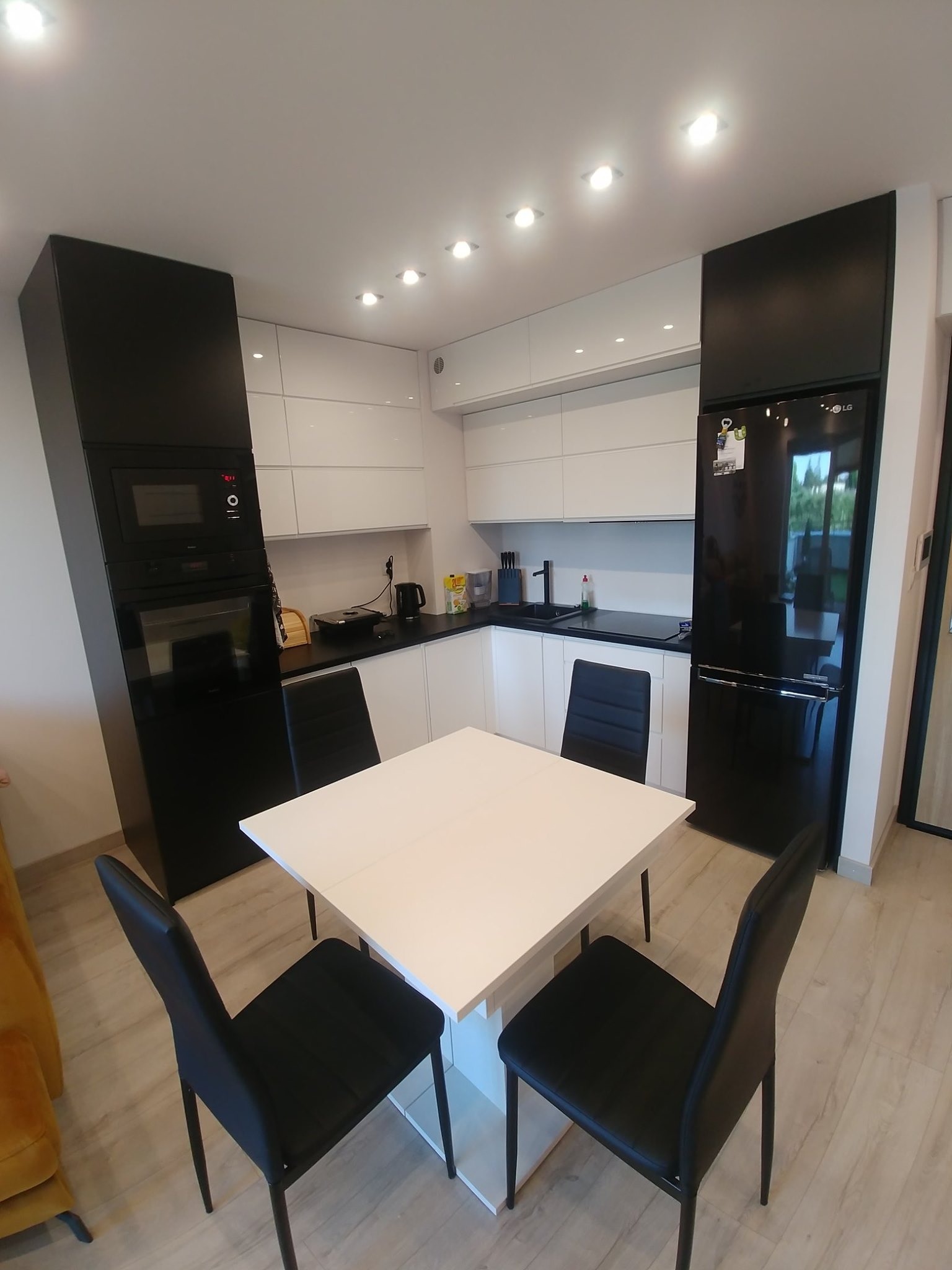 Nowoczesna kuchnia z białymi szafkami, czarnymi elementami i białym stołem otoczonym czarnymi krzesłami, oświetlona punktowymi lampami sufitowymi.