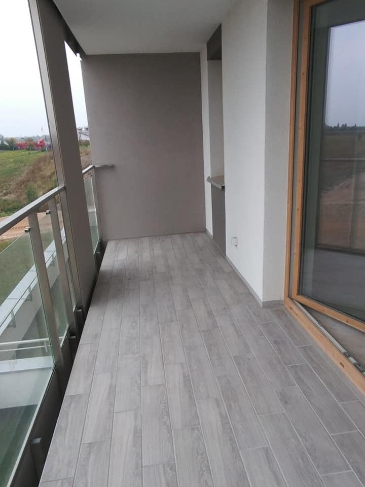 Nowoczesny balkon z szarą podłogą imitującą drewno, przeszklona balustrada, widok na zewnątrz w pochmurny dzień.
