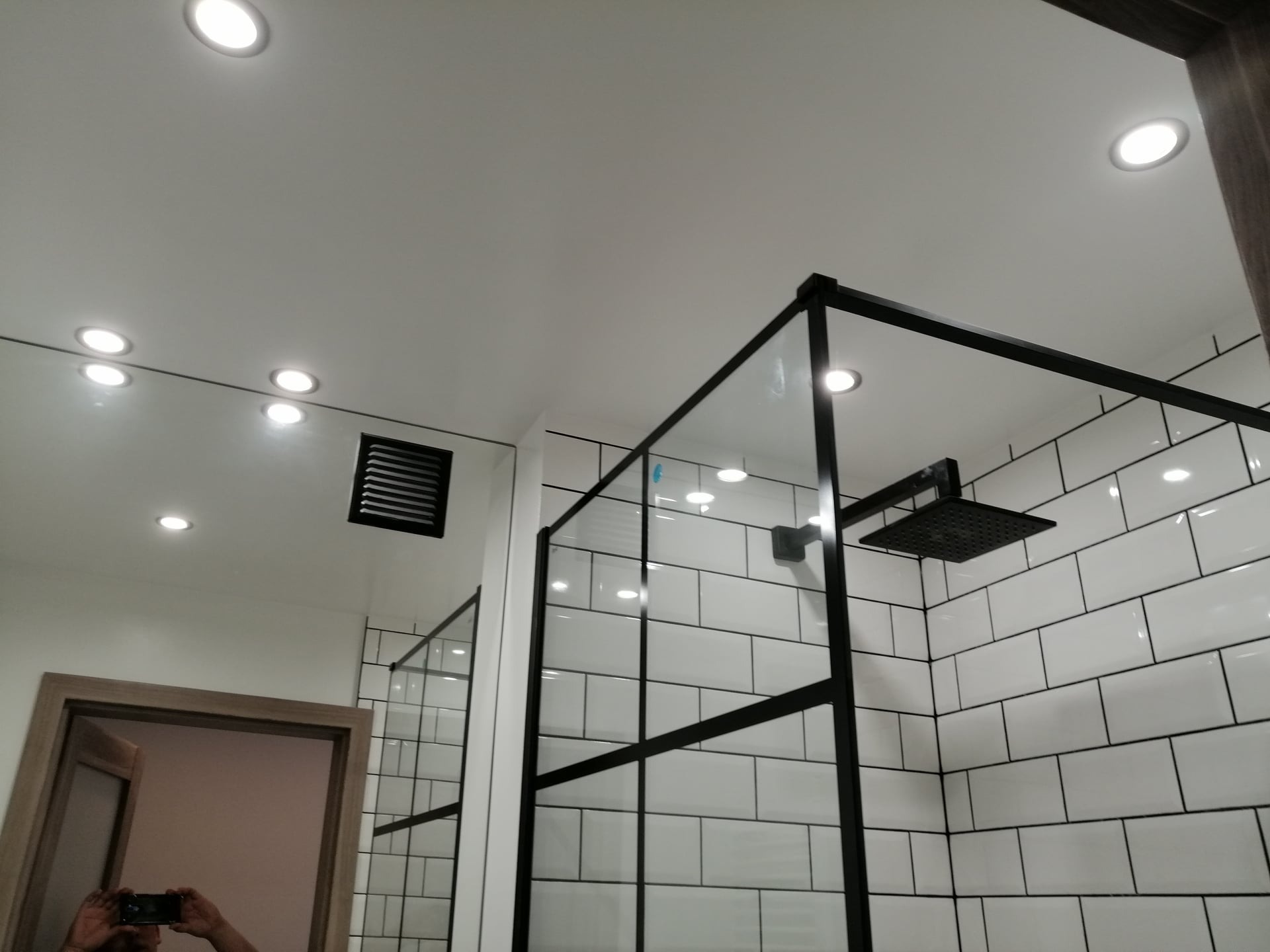 Nowoczesna kabina prysznicowa typu walk-in z czarnymi profilami, białe płytki ułożone w cegiełkę, oświetlenie punktowe w suficie, widoczne odbicie fotografa w lustrze.