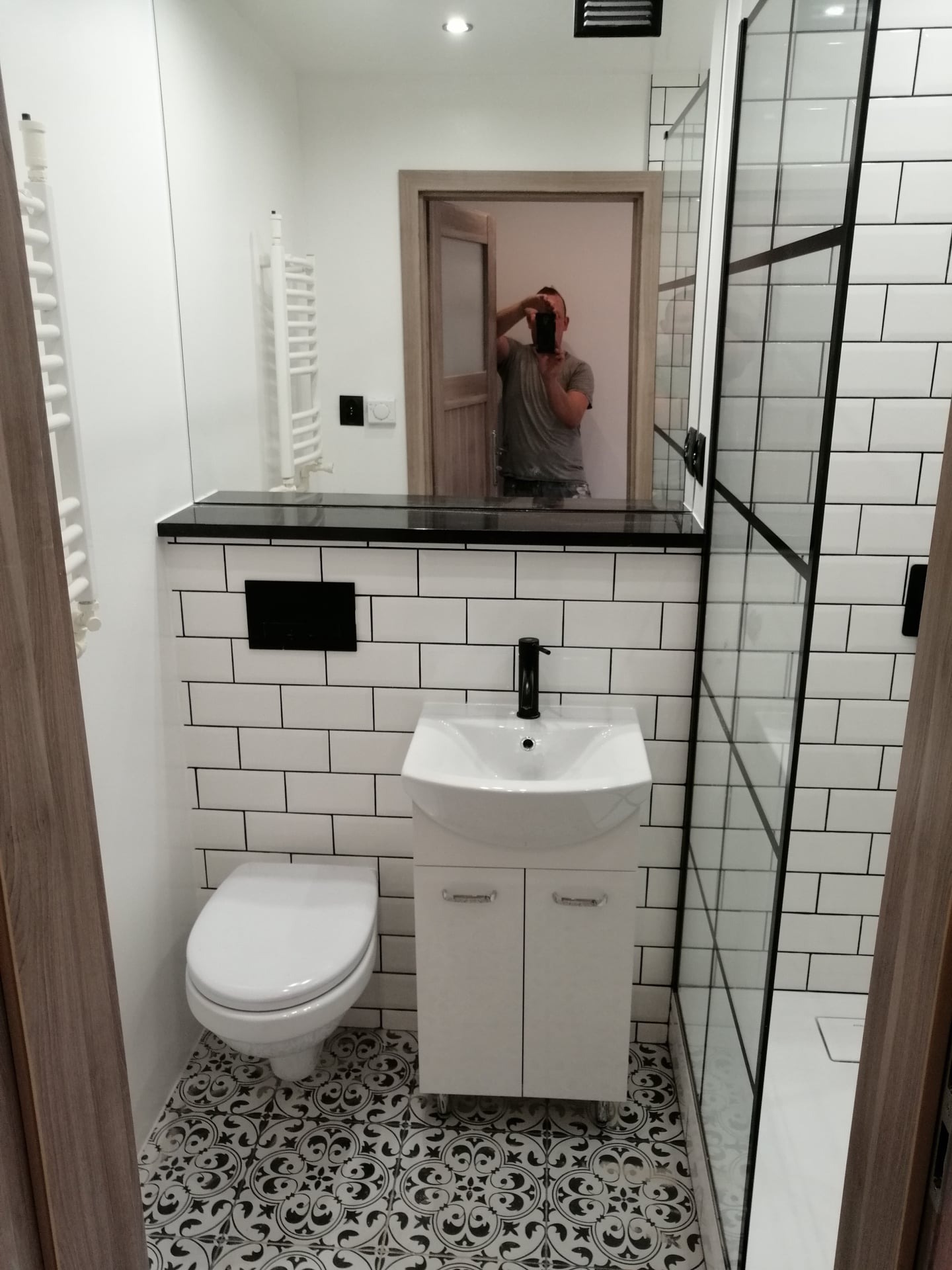 Nowoczesna łazienka z białą armaturą, czarnymi akcentami i wzorzystą podłogą; widoczne płytki typu metro, kabina prysznicowa z czarnymi ramami i odbicie fotografa w lustrze.