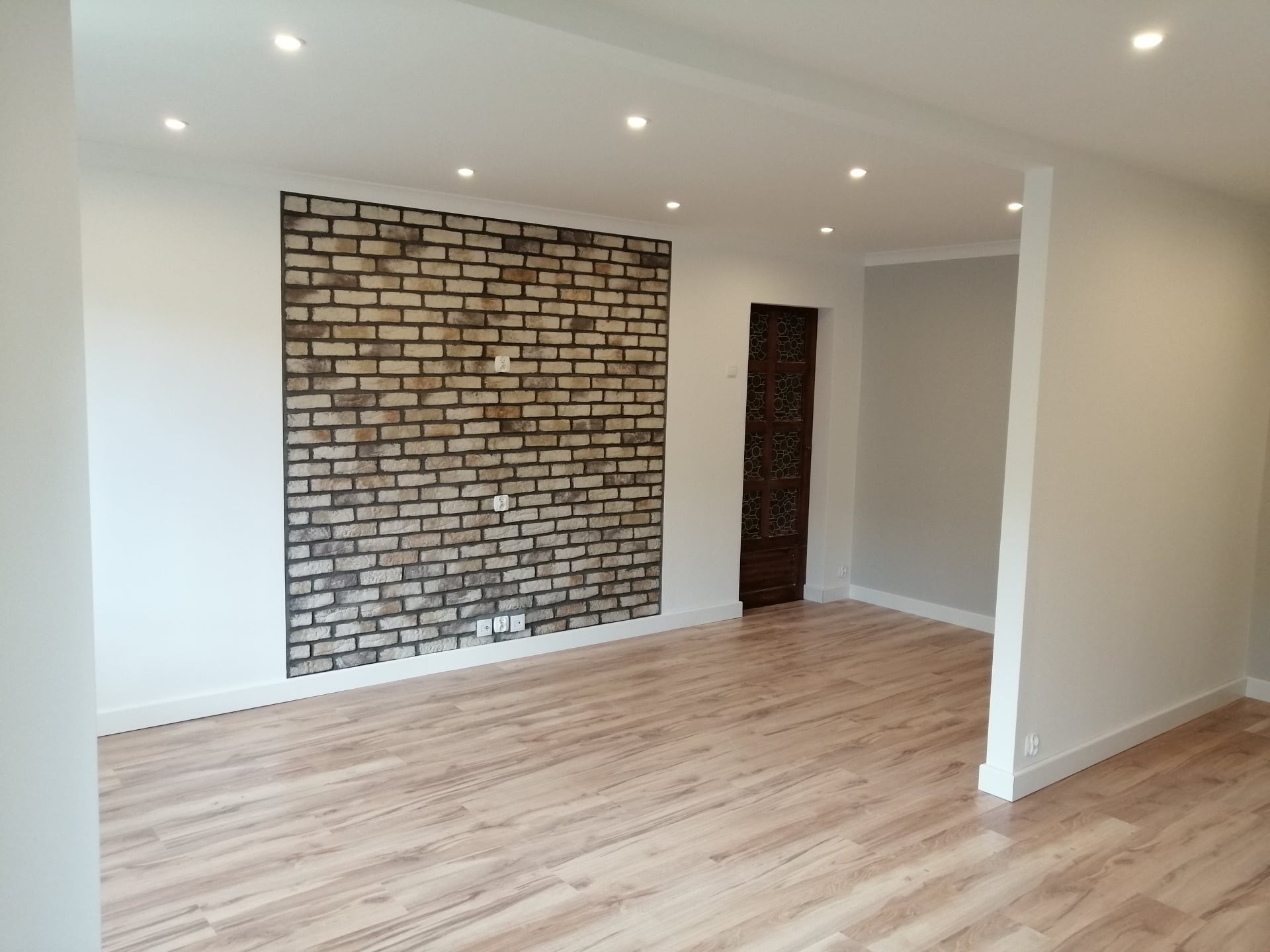 Wykończone wnętrze pokoju z panelami podłogowymi imitującymi drewno i ścianą akcentową z cegły dekoracyjnej, z widocznymi gniazdkami elektrycznymi, oświetleniem sufitowym i fragmentem drewnianych...