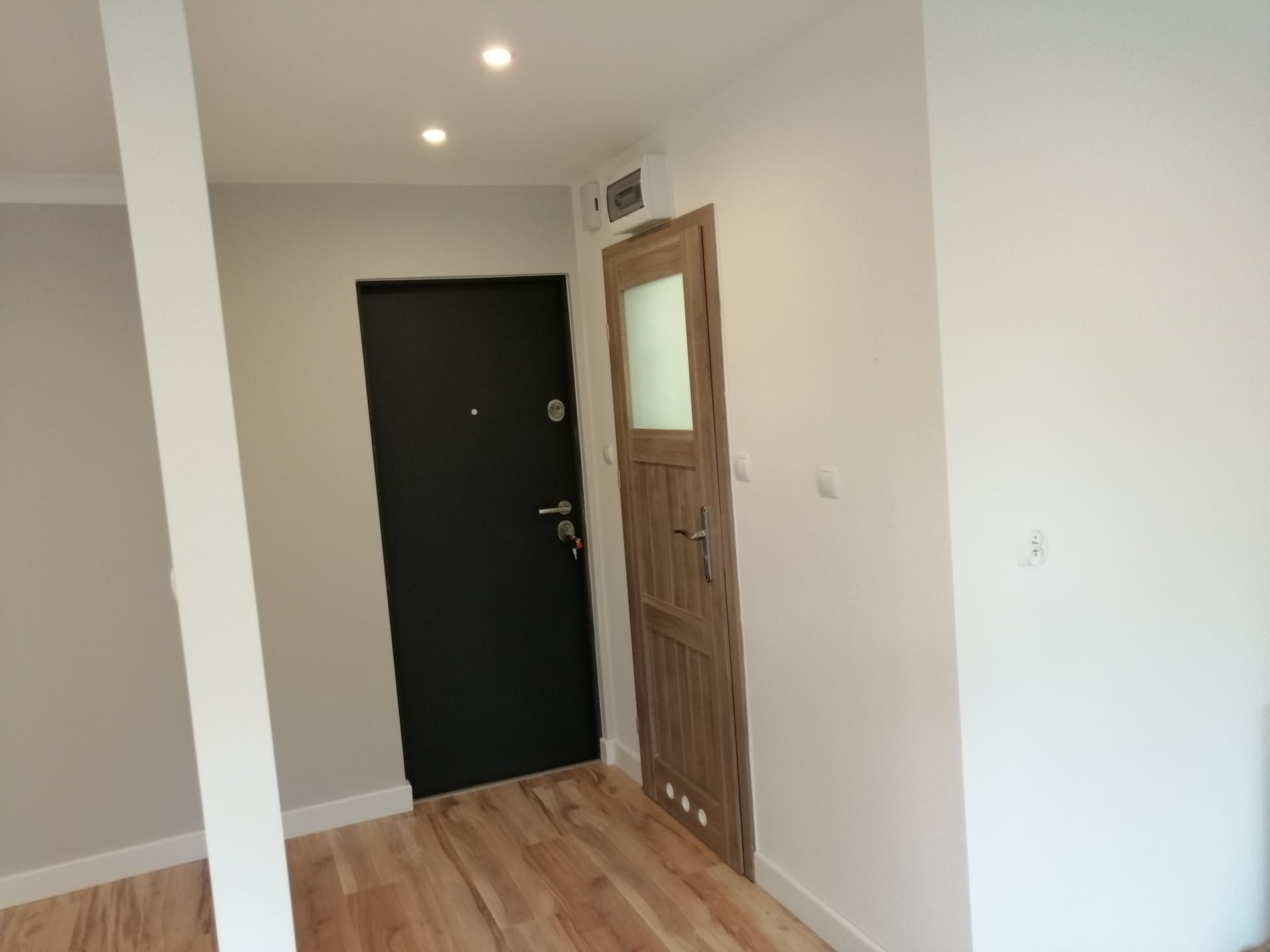 Korytarz po remoncie z dwiema nowymi drzwiami: ciemnymi antywłamaniowymi i drewnianymi z przeszkleniem, jasne ściany i podłoga z paneli.