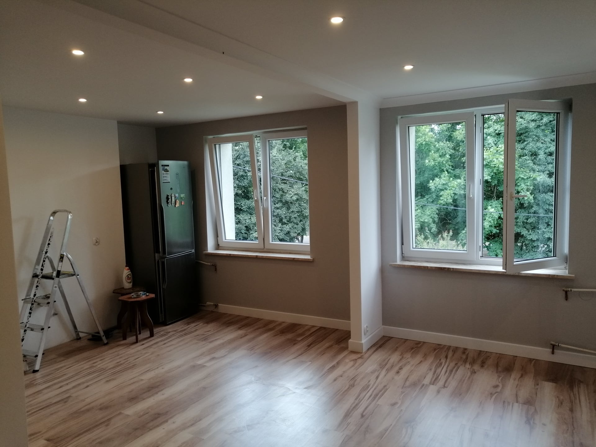 Jasne, przestronne wnętrze po remoncie z drewnopodobną podłogą, dwoma oknami z widokiem na zieleń, aluminiową drabiną opartą o ścianę obok czarnej lodówki, oraz z wbudowanymi punktowymi lampami...