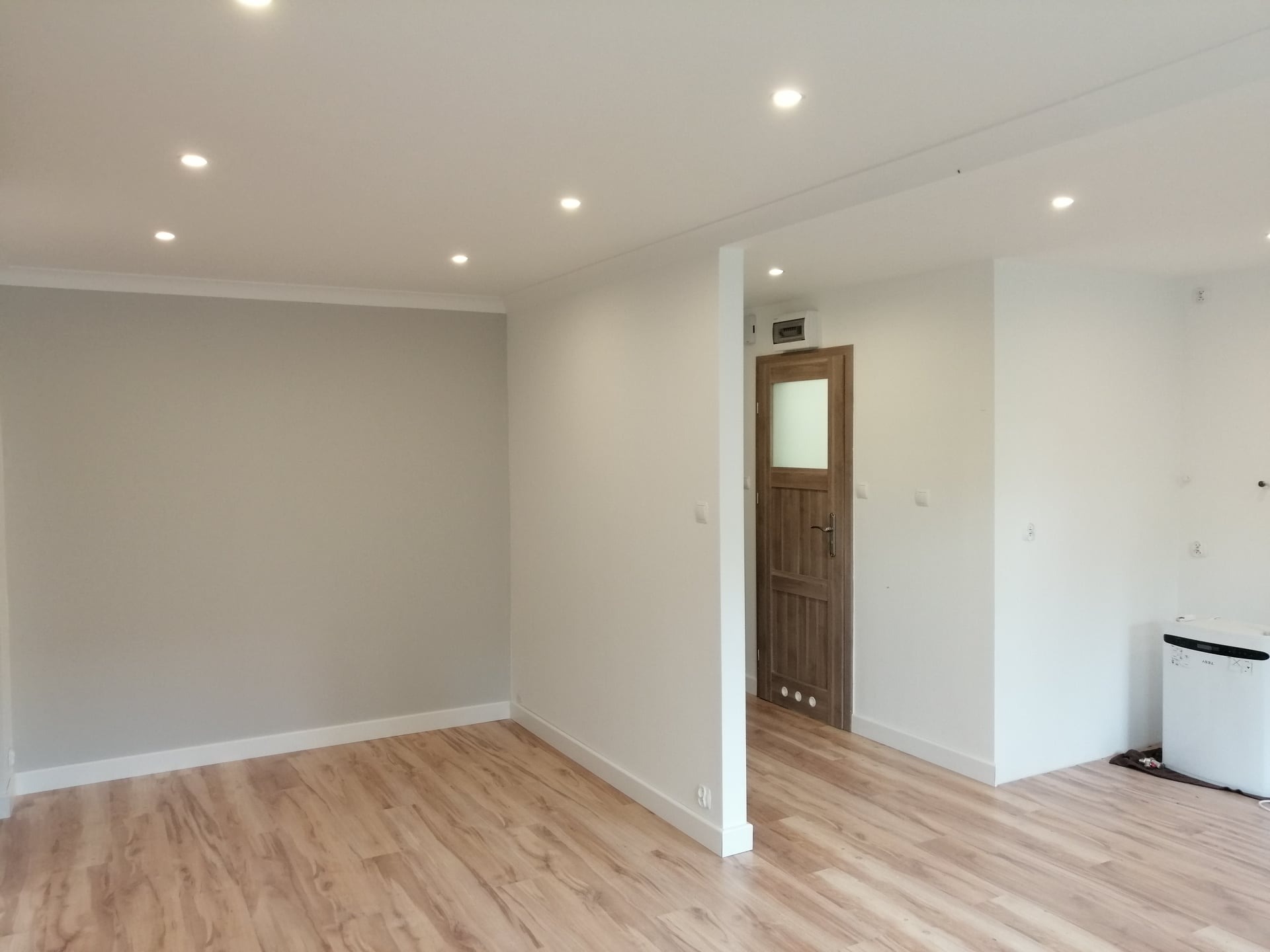 Jasne, przestronne wnętrze po remoncie w Suwałkach: ściany pomalowane na szaro i biało, drewnopodobna podłoga, białe listwy przypodłogowe, drewniane drzwi z szybą i punktowe oświetlenie sufitowe.