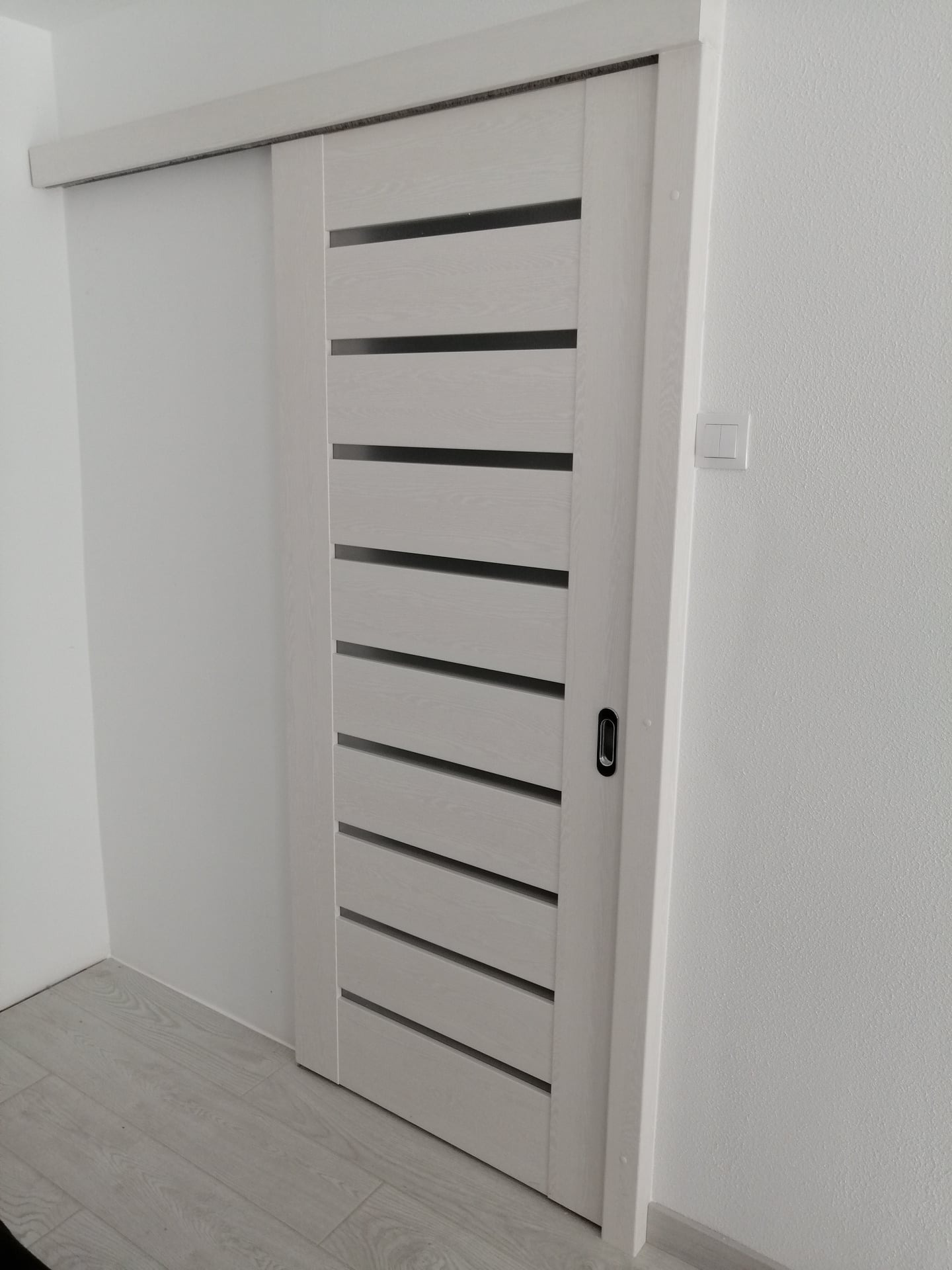 Białe przesuwne drzwi z poziomymi, ciemnymi paskami, zamontowane w białej ścianie, widoczny fragment podłogi z jasnymi panelami.