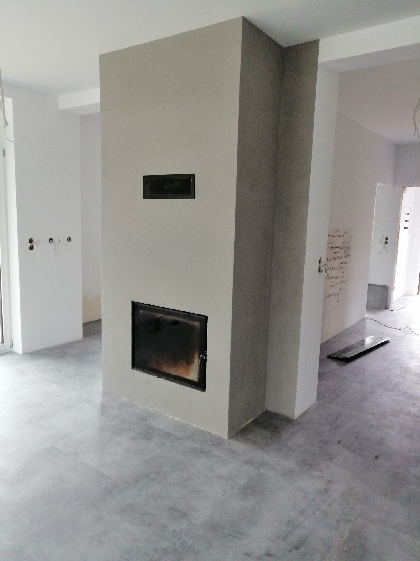 Surowy stan wnętrza z obudowanym kominkiem w odcieniach szarości, widoczne przygotowania do wykończenia ścian i podłóg, brak listew przypodłogowych, instalacja elektryczna bez osprzętu, płyta...