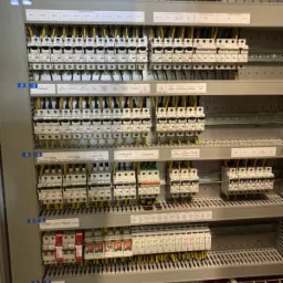 Rozdzielnica elektryczna z licznymi zabezpieczeniami i okablowaniem, widoczne etykiety opisowe na poszczególnych elementach.