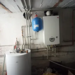 Montaż kotła grzewczego Termet i niebieskiego zbiornika wyrównawczego w piwnicy z widocznymi rurami instalacji centralnego ogrzewania i narzędziami na górze bojlera.