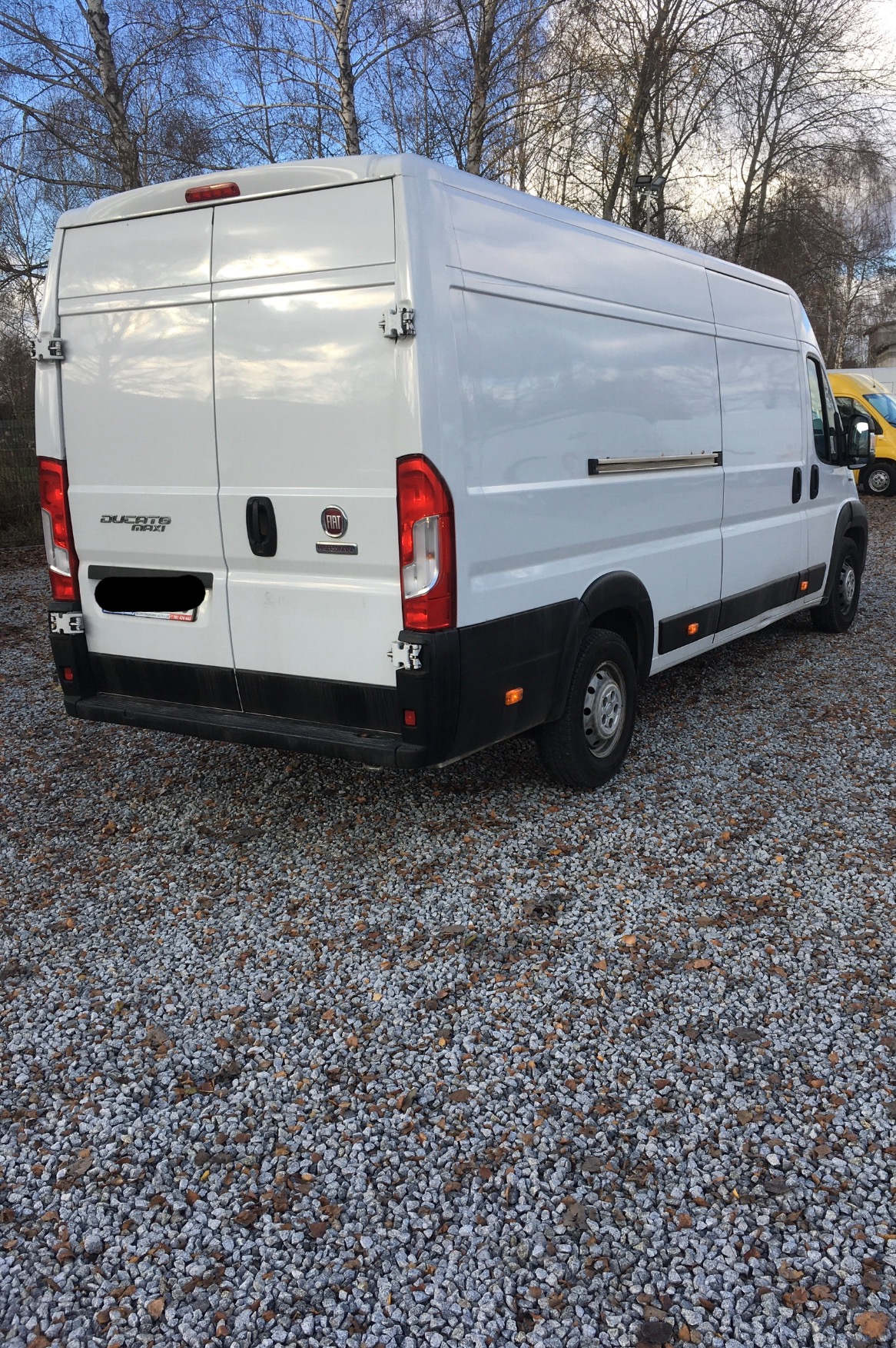 Biały furgon Fiat Ducato Maxi zaparkowany na żwirowym podłożu, widok z tyłu i boku, z widocznymi detalami jak logo i oznaczenia modelu.