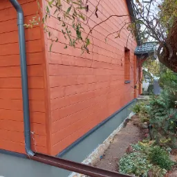 Boczny widok domu z drewnianą elewacją pomalowaną na pomarańczowo, szara podmurówka, system rynnowy i fragment ogrodu z roślinnością.