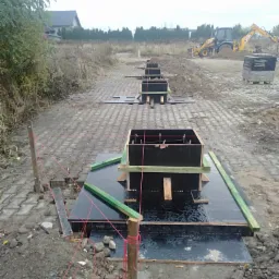 Zalane szalunki pod fundamenty w trakcie budowy, widoczny wykop i koparka w tle, teren przygotowany pod wylewkę.