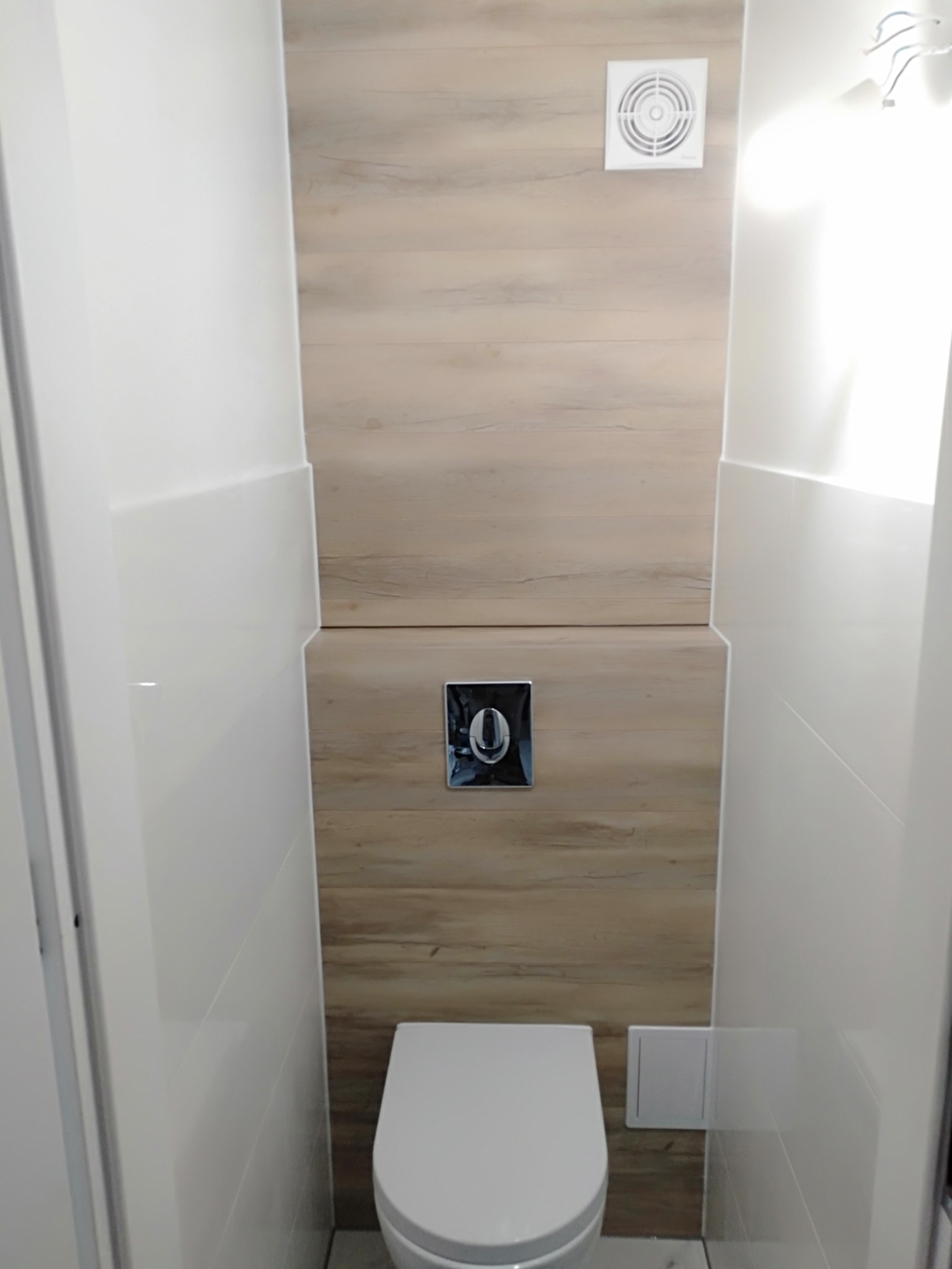 Wąska toaleta z zabudową z paneli imitujących drewno, podwieszana miska ustępowa i chromowany przycisk spłukujący, wentylator w górnej części ściany.