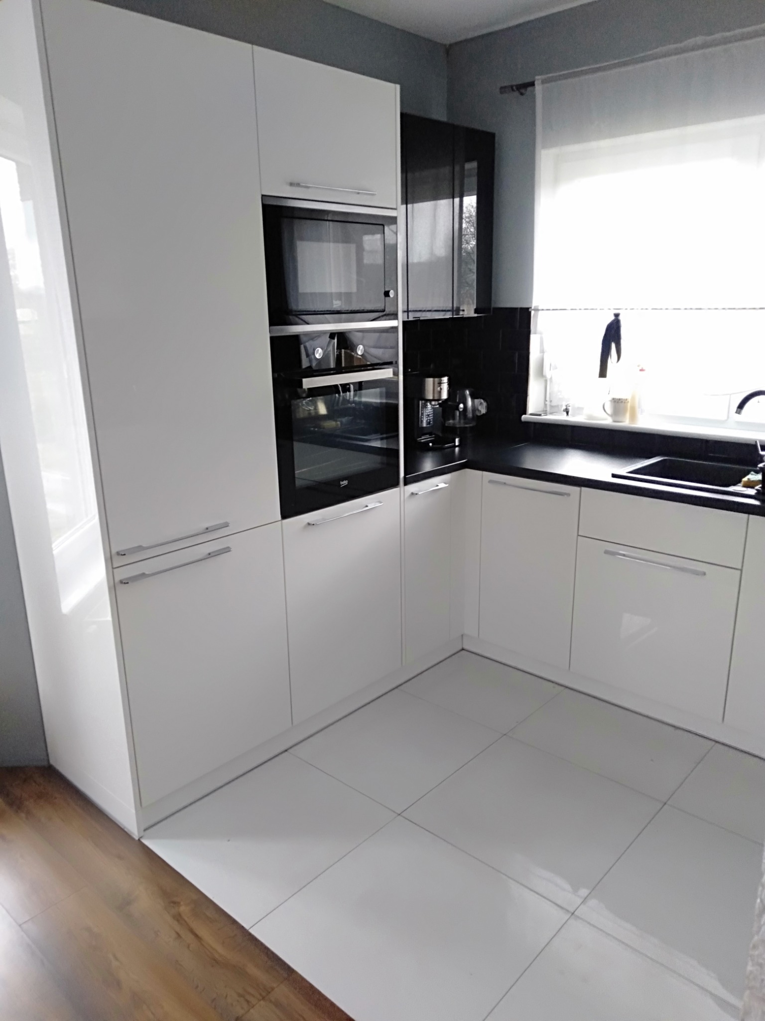 Nowoczesna kuchnia z białymi, błyszczącymi szafkami, czarnym blatem i płytkami, z widocznymi urządzeniami AGD w zabudowie, takimi jak piekarnik i mikrofalówka marki Beko.