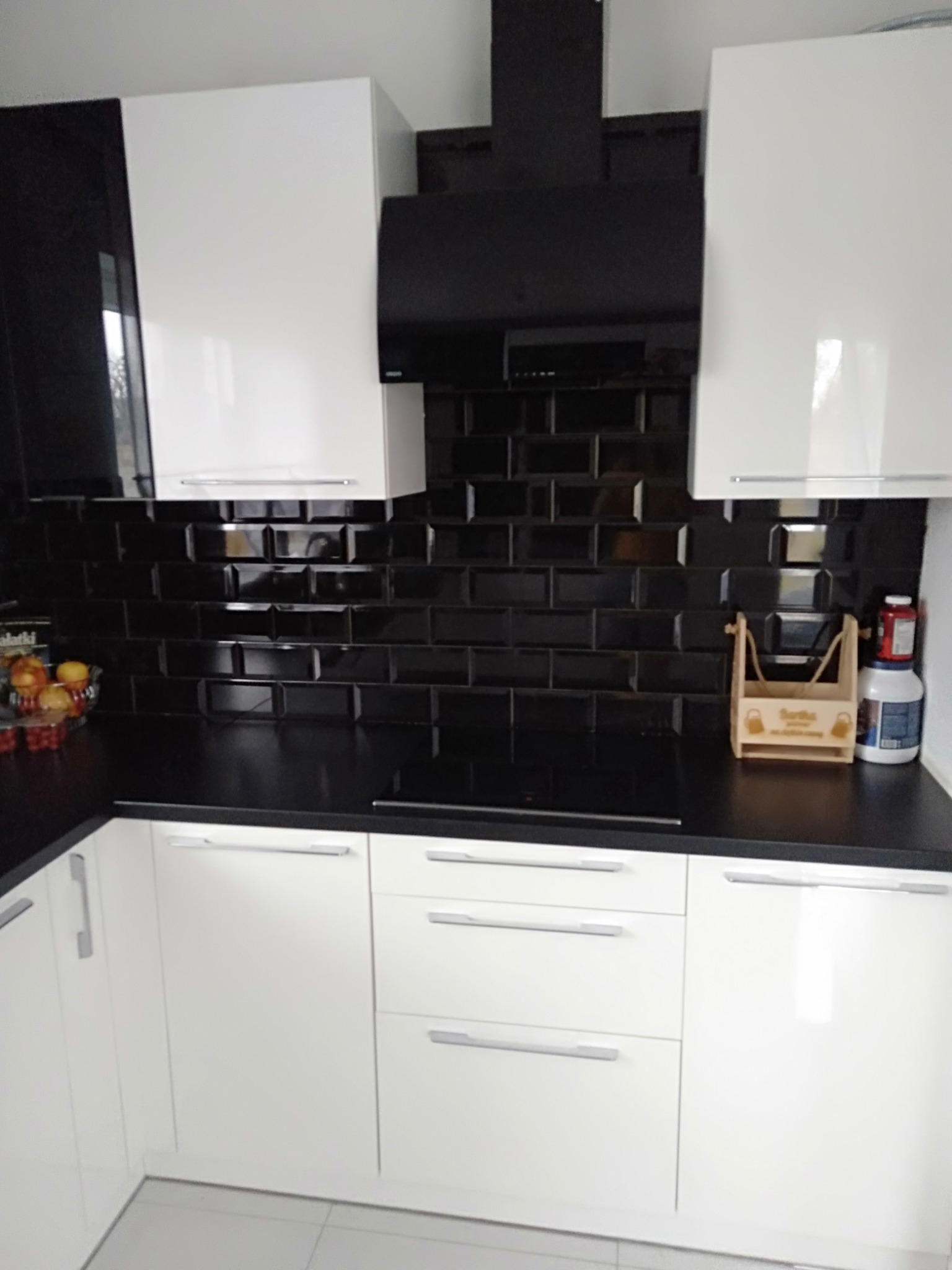 Nowoczesna kuchnia z białymi, błyszczącymi szafkami i czarnym blatem oraz czarnymi płytkami ściennymi w stylu metro, drewniany organizer na blacie.