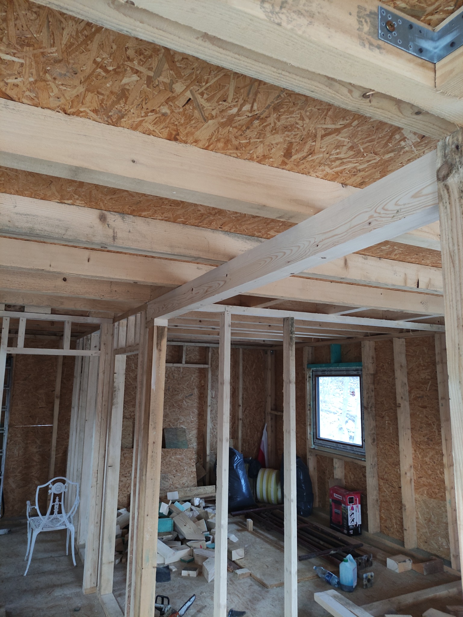 Wnętrze w trakcie budowy: surowa konstrukcja szkieletowa z płyt OSB, widoczne drewniane belki stropowe, białe krzesło i narzędzia budowlane wewnątrz pomieszczenia.