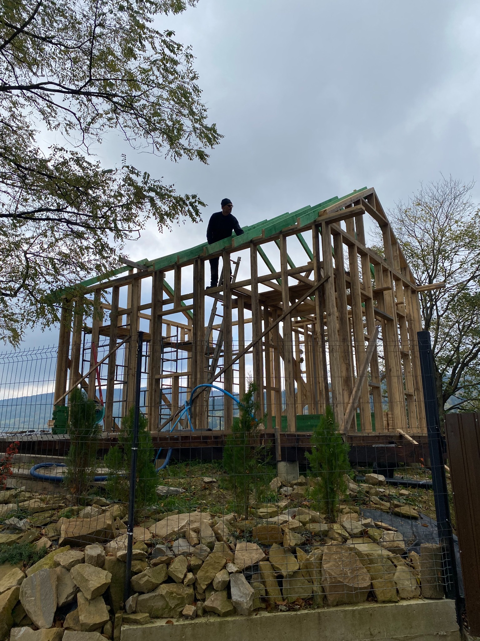 Konstrukcja drewniana domu w trakcie budowy, widoczny pracownik na dachu, szkielet budynku z zielonymi elementami, otoczenie górskie z drzewami i ogrodzeniem.