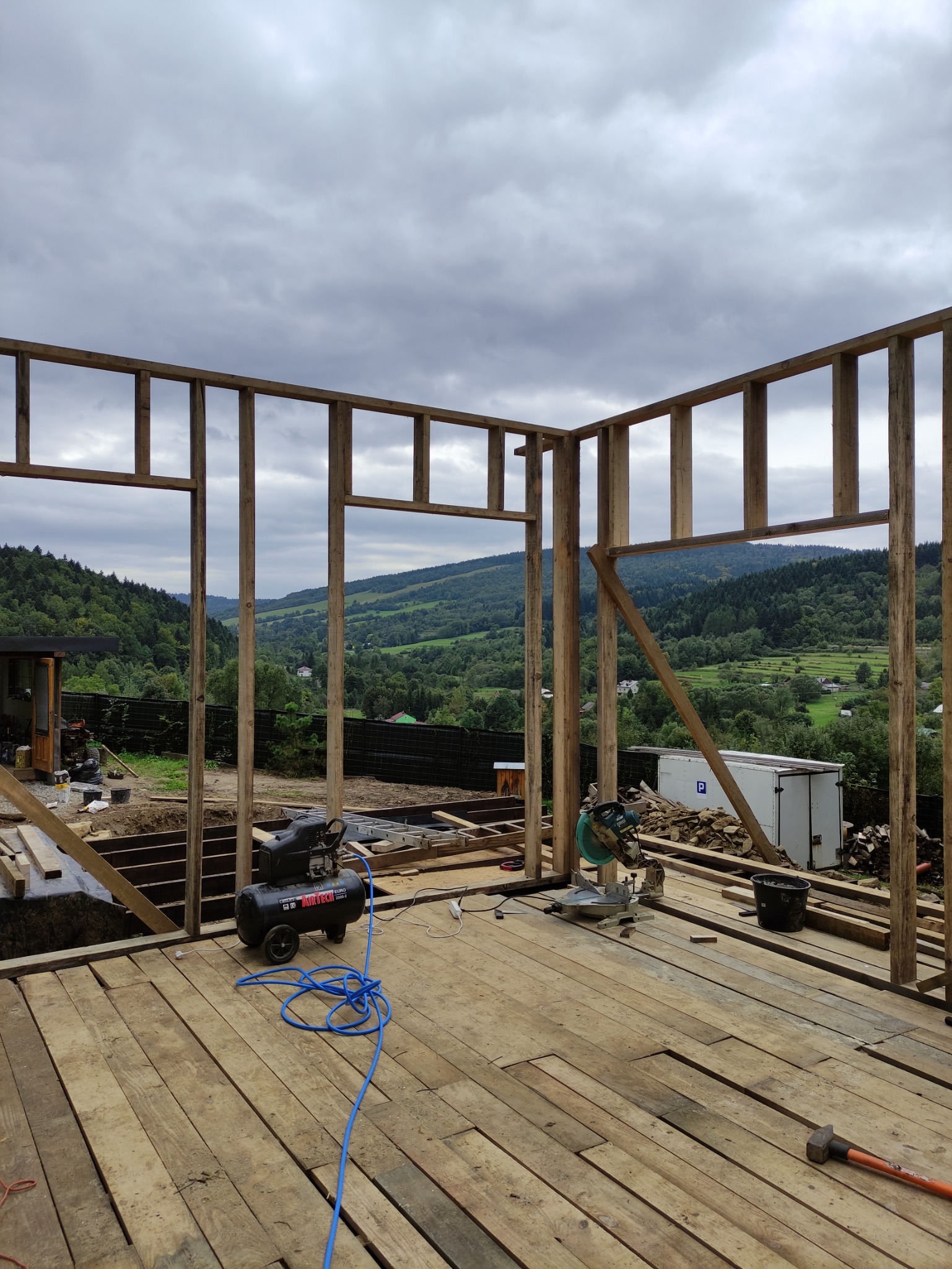 Wnętrze domu szkieletowego w trakcie budowy, widoczny kompresor, piła ukośnica i panorama gór w tle.