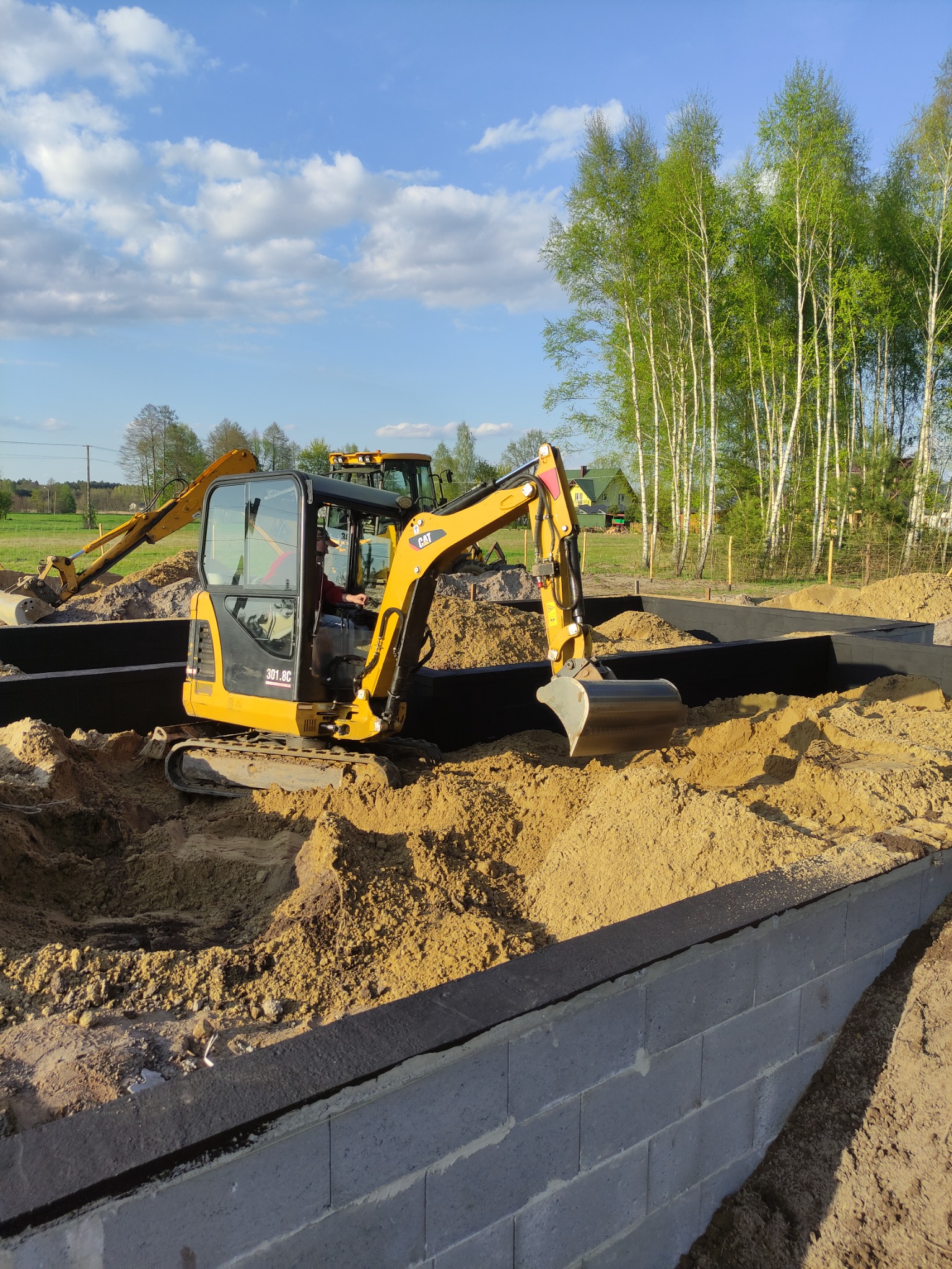 Żółta koparka CAT 301.8C w wykopie fundamentowym z bloczków betonowych, wypełnionym piaskiem, w tle druga koparka i brzozowy lasek.