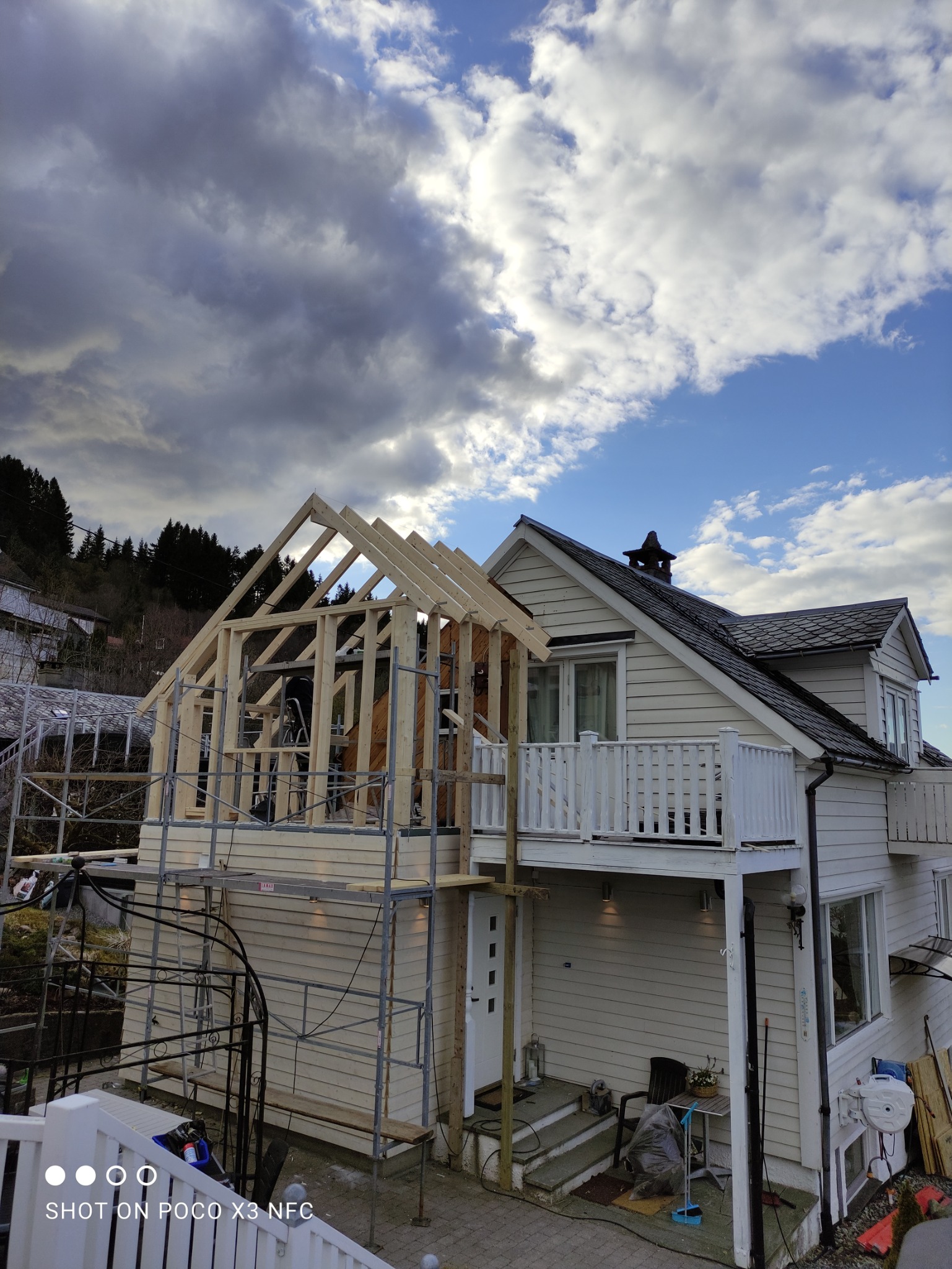 Wznoszenie drewnianej konstrukcji domu, widoczne rusztowania i fragment istniejącego budynku z balkonem, pochmurne niebo.