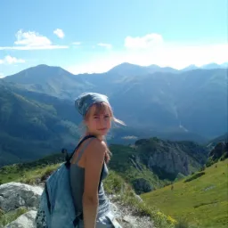 Młoda kobieta z plecakiem i chustą na głowie siedzi na skalistym szczycie góry, patrząc w obiektyw, z rozległym widokiem na góry i błękitne niebo w tle.