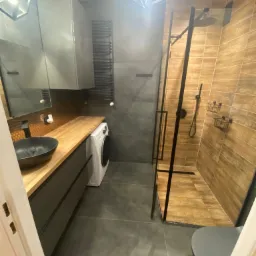 Nowoczesna łazienka z drewnianymi akcentami: blat z jasnego drewna, umywalka nablatowa w kolorze czarnym, kabina prysznicowa z czarnymi ramami i płytkami imitującymi drewno, pralka wkomponowana...