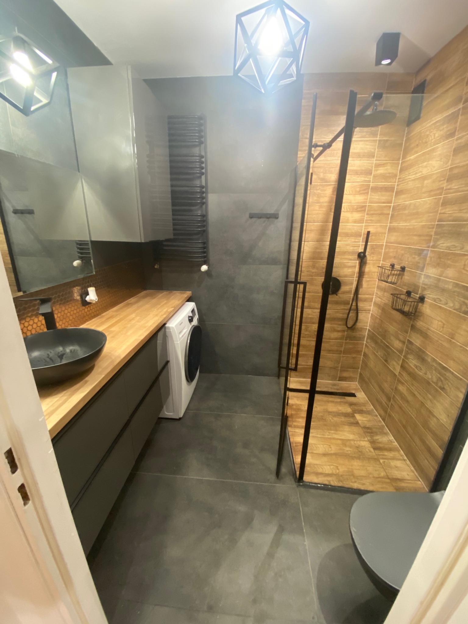 Nowoczesna łazienka z drewnianymi akcentami: blat z jasnego drewna, umywalka nablatowa w kolorze czarnym, kabina prysznicowa z czarnymi ramami i płytkami imitującymi drewno, pralka wkomponowana...