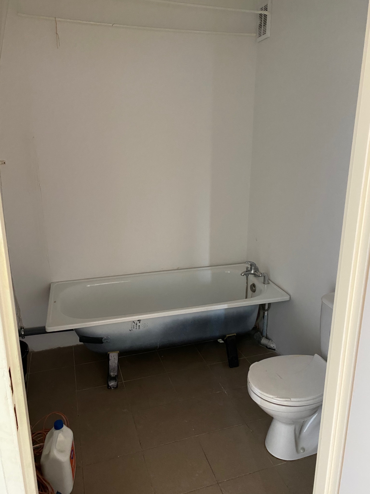 Łazienka przed remontem z białą wanną na nóżkach, brązowymi płytkami na podłodze i toaletą z białym sedesem; widoczny biały pojemnik z płynem do czyszczenia.