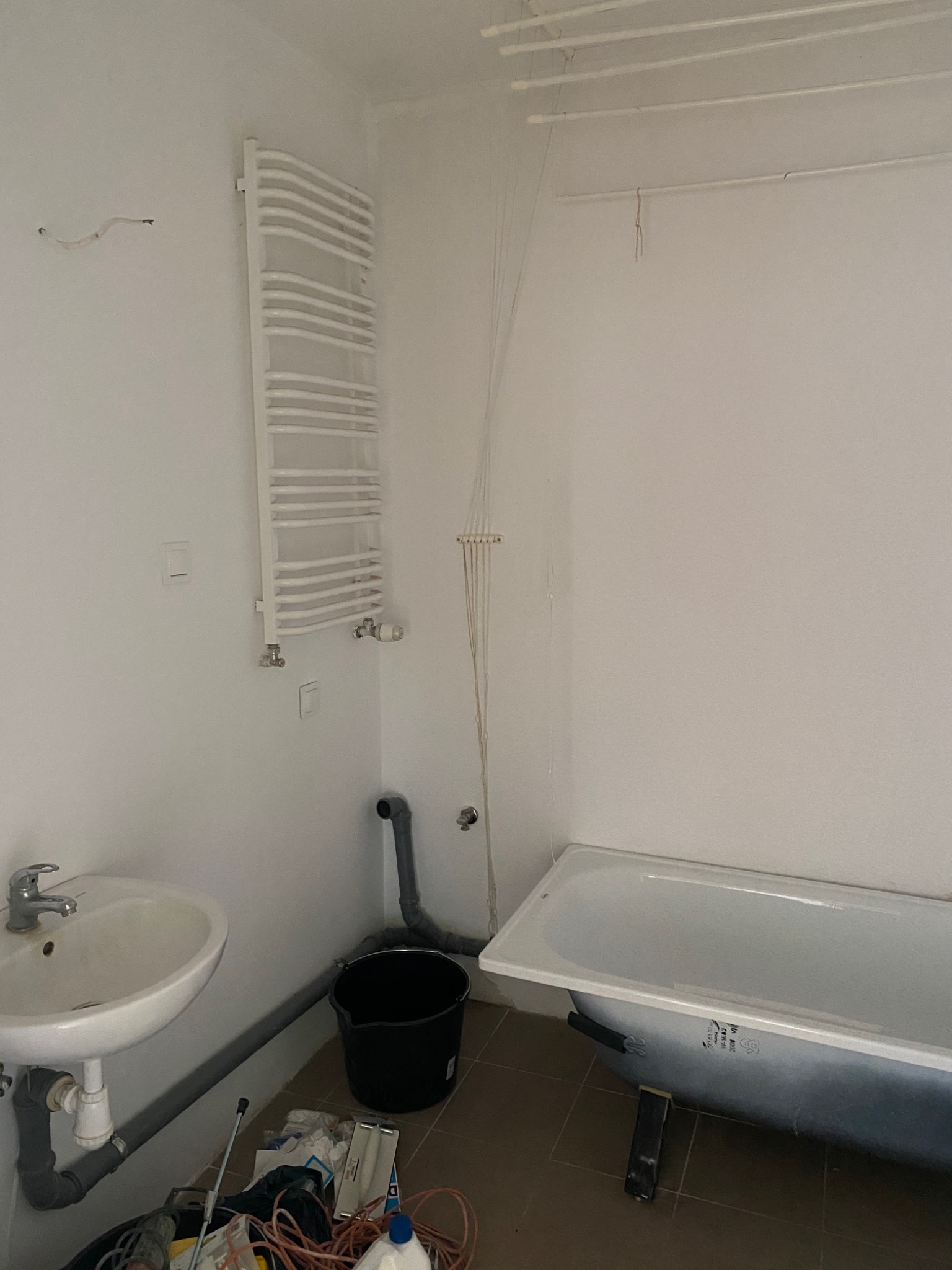 Łazienka w trakcie remontu: widoczna wanna, umywalka, grzejnik drabinkowy, pranie pod sufitem, wiadro i narzędzia na podłodze.