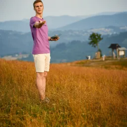 Młody mężczyzna w fioletowej koszulce kontroluje drona na tle górskiego krajobrazu, z widocznym kontrolerem i wyciągniętą dłonią.