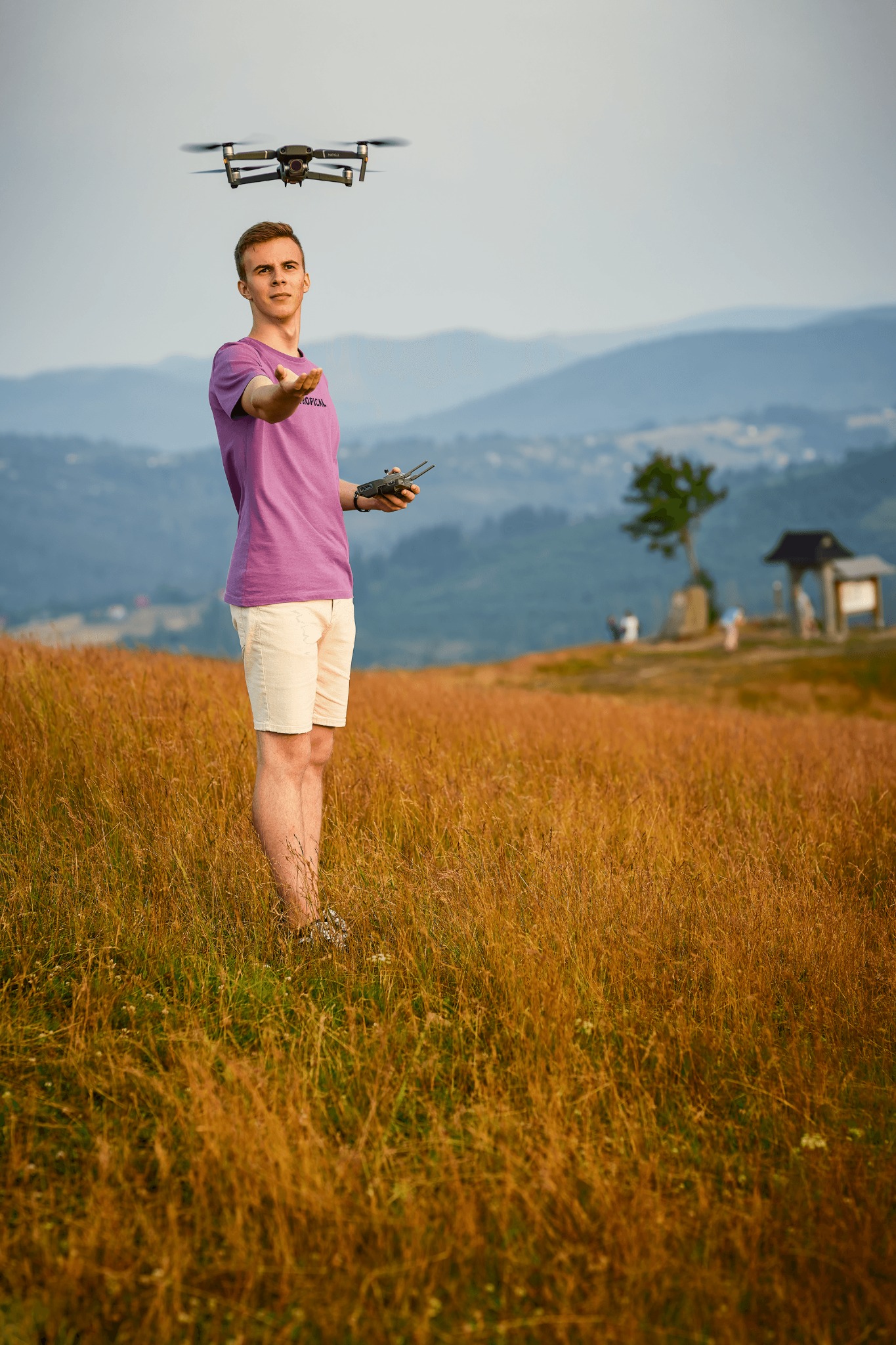 Młody mężczyzna w fioletowej koszulce kontroluje drona na tle górskiego krajobrazu, z widocznym kontrolerem i wyciągniętą dłonią.
