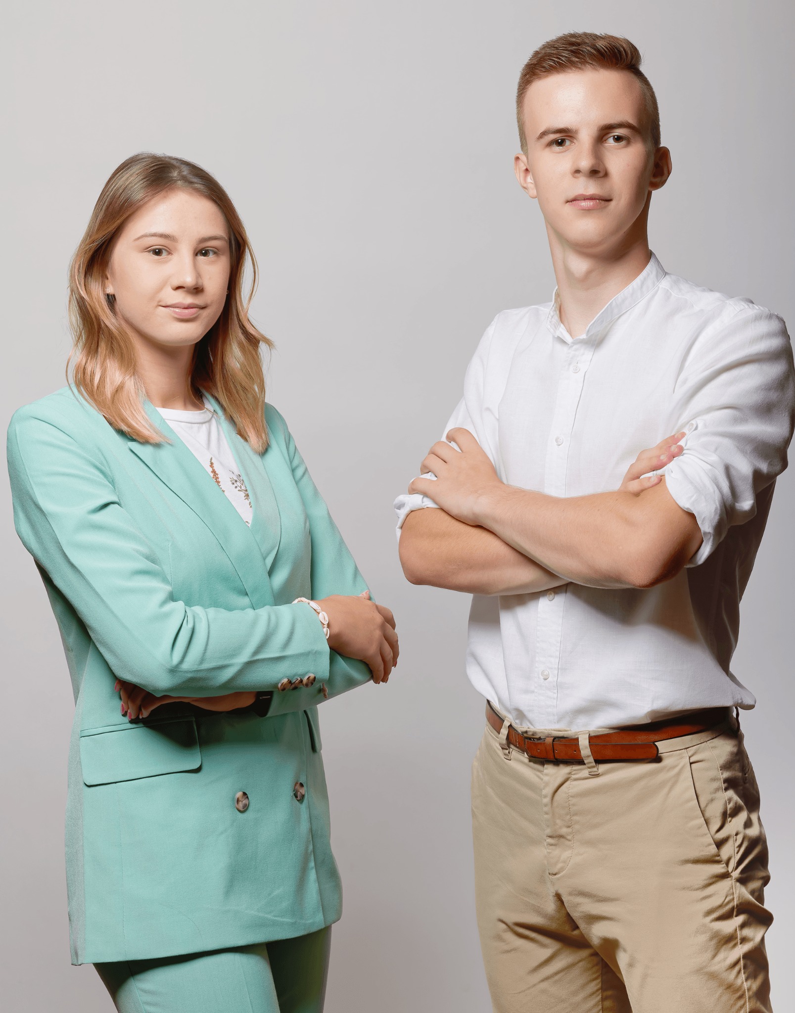 Dwoje młodych ludzi, kobieta w miętowym garniturze i mężczyzna w białej koszuli i beżowych spodniach, stoją na jasnym tle ze skrzyżowanymi ramionami, patrząc w kamerę.