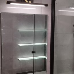 CZECH PRO-Bud - Nowoczesna łazienka z szarymi płytkami imitującymi marmur, podświetlana szafka z lustrem i czarna lampa nad nią, fragment kabiny prysznicowej z czarną ramą.
