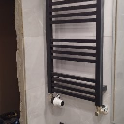 CZECH PRO-Bud - Czarny grzejnik łazienkowy drabinkowy zamontowany na ścianie z płytek imitujących marmur, widoczne przyłącza i regulator temperatury, obok gniazdko elektryczne z podświetlanym włącznikiem...