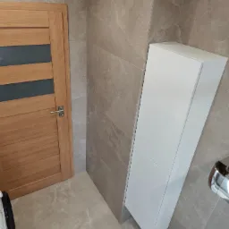 Wykończona łazienka z beżowymi płytkami imitującymi marmur, drewnianymi drzwiami z przeszkleniem i białą, wąską szafką. Nowoczesny design.