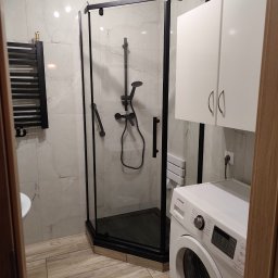 CZECH PRO-Bud - Nowoczesna kabina prysznicowa z czarnymi profilami, marmurowe płytki ścienne, drewnopodobna podłoga, biała pralka i szafka w tle. Całość w jasnej, przestronnej łazience.