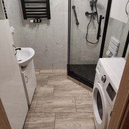 CZECH PRO-Bud - Nowoczesna łazienka z kabiną prysznicową typu walk-in, drewnopodobną podłogą, umywalką z szafką i czarnym grzejnikiem drabinkowym.