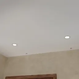 Biały sufit napinany z trzema okrągłymi lampami LED, ściany wykończone w beżowym tynku dekoracyjnym.