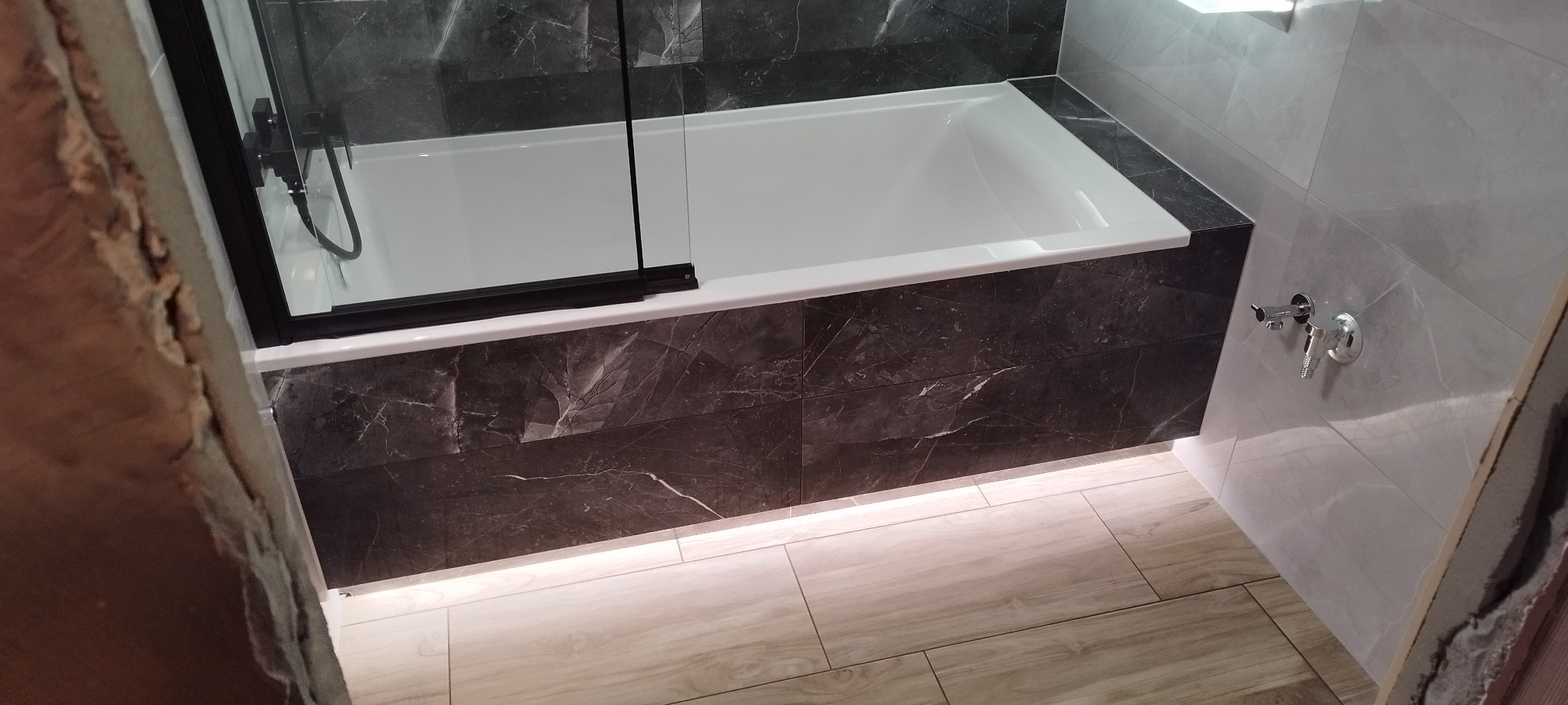 Nowoczesna łazienka z wanną obudowaną czarnymi płytkami imitującymi marmur, podświetlenie LED u dołu obudowy, szklany parawan nawannowy z czarną ramą, jasne płytki podłogowe imitujące drewno...