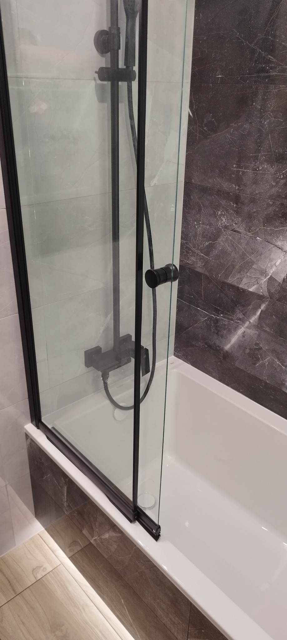Kabina prysznicowa z czarnym osprzętem, zamontowana na wannie, widok na szarą marmurową ścianę i beżową podłogę z płytek.