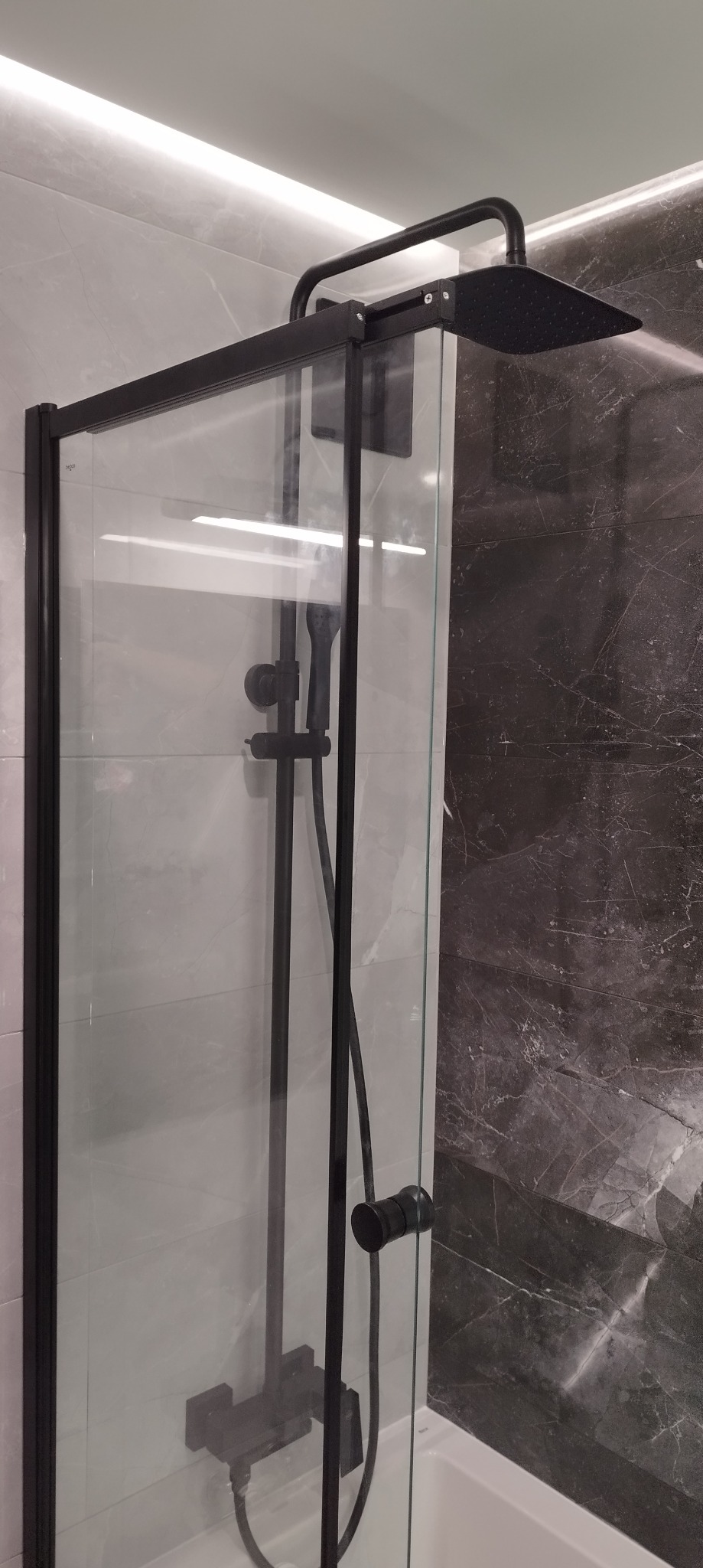 Szklana kabina prysznicowa z czarnymi profilami i deszczownicą na tle marmurowych i jasnych płytek ceramicznych, widoczny fragment wanny.