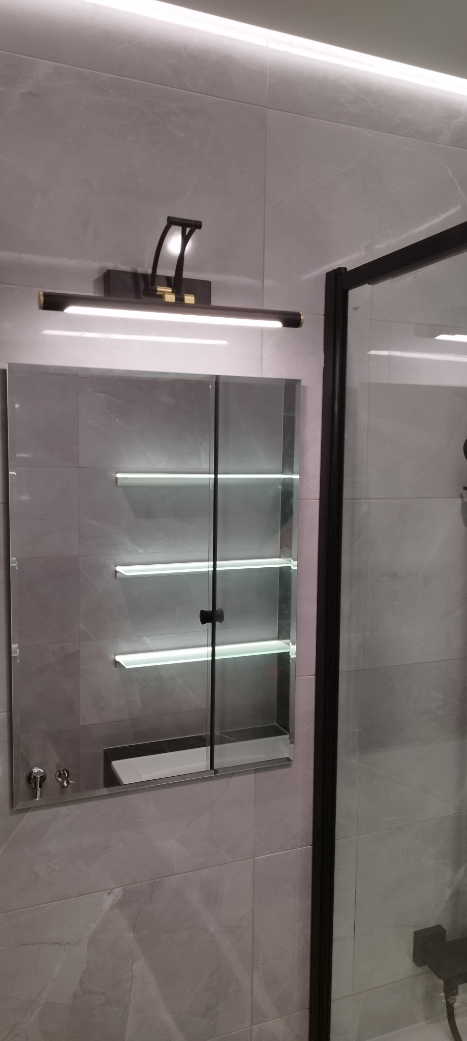 Nowoczesna łazienka z szarymi płytkami imitującymi marmur, podświetlana szafka z lustrem i czarna lampa nad nią, fragment kabiny prysznicowej z czarną ramą.