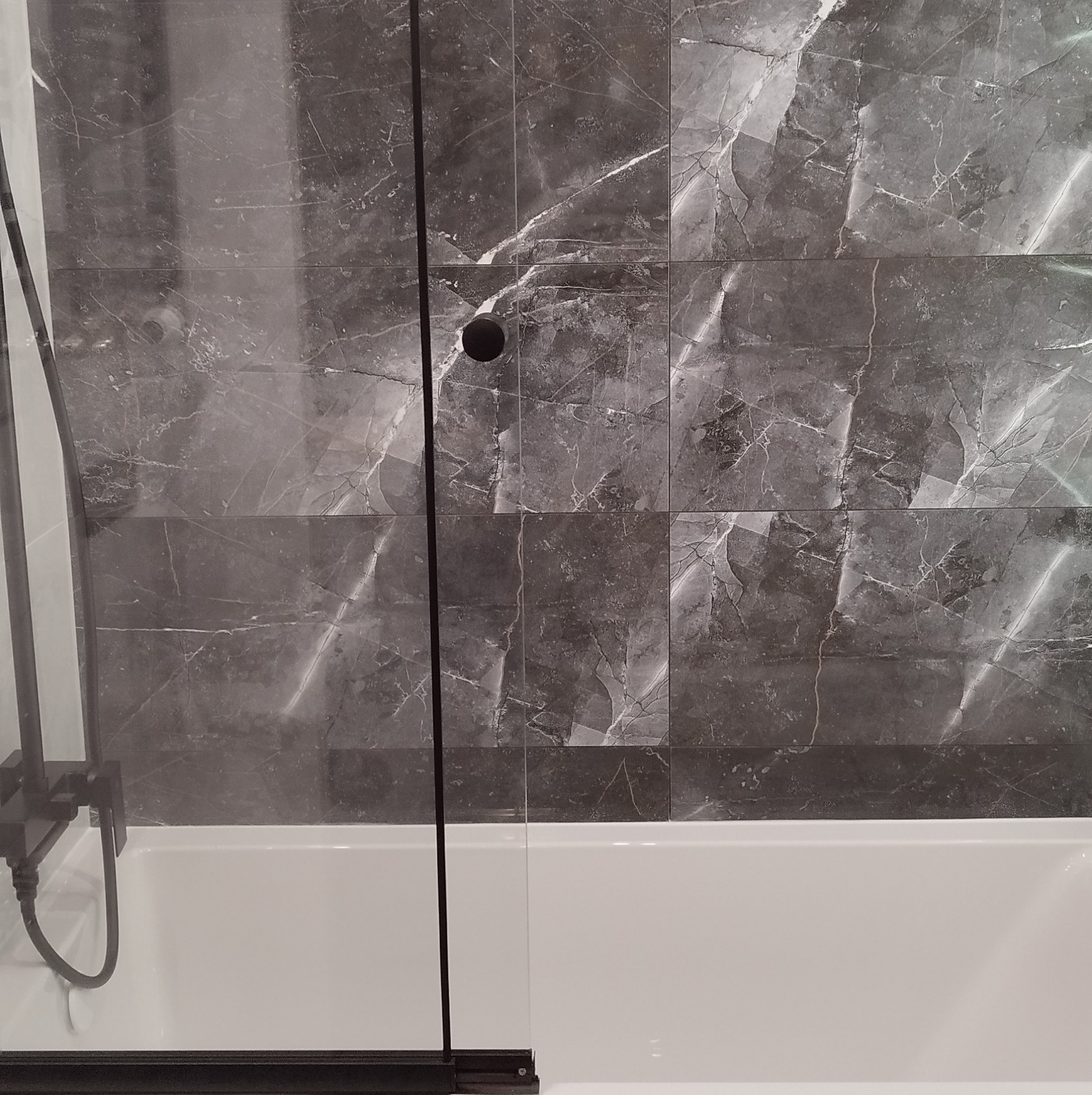 Szare płytki imitujące marmur na ścianie nad białą wanną, częściowo zasłonięte czarną ramą szklanego parawanu nawannowego i czarną słuchawką prysznicową.